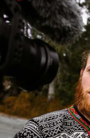 A bearded man records a video outdoors using a professional camera, showcasing selective focus and vlogging setup.
