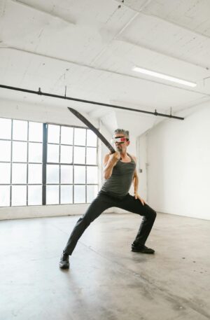 Adult man posing with a sword and VR headset in a minimalistic studio.